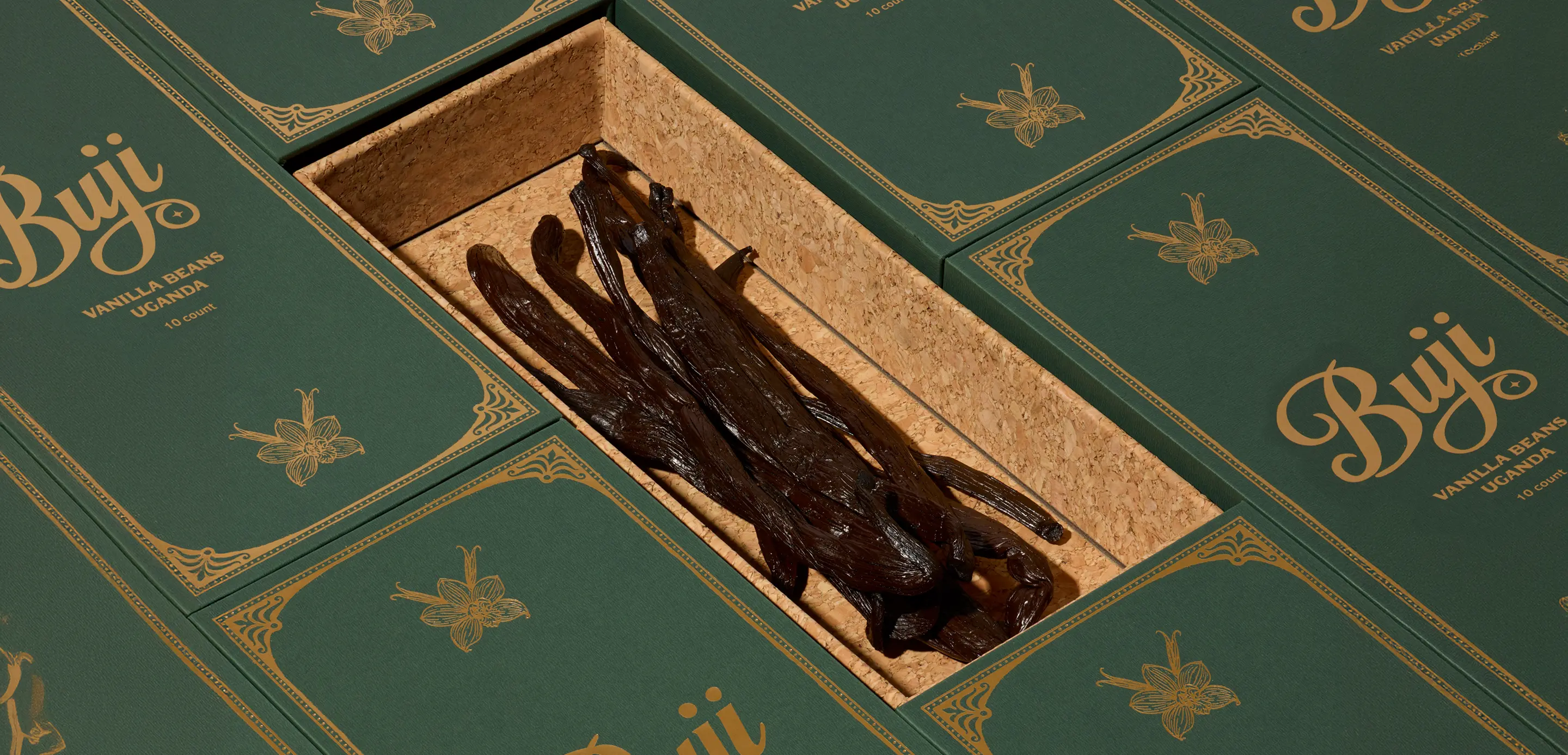 Dark vanilla beans in an open cork box surrounded by green boxes labeled Buji vanilla beans Uganda.