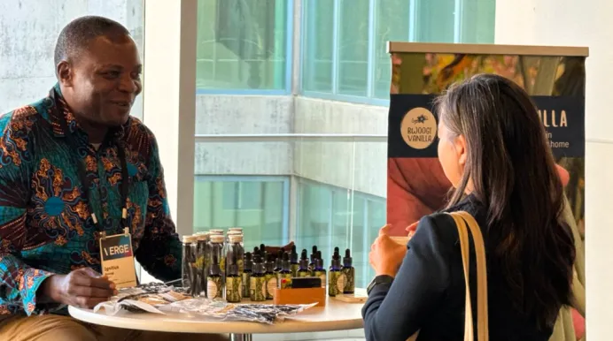 Man and woman conversing at a table with vanilla product bottles and samples at an indoor event.