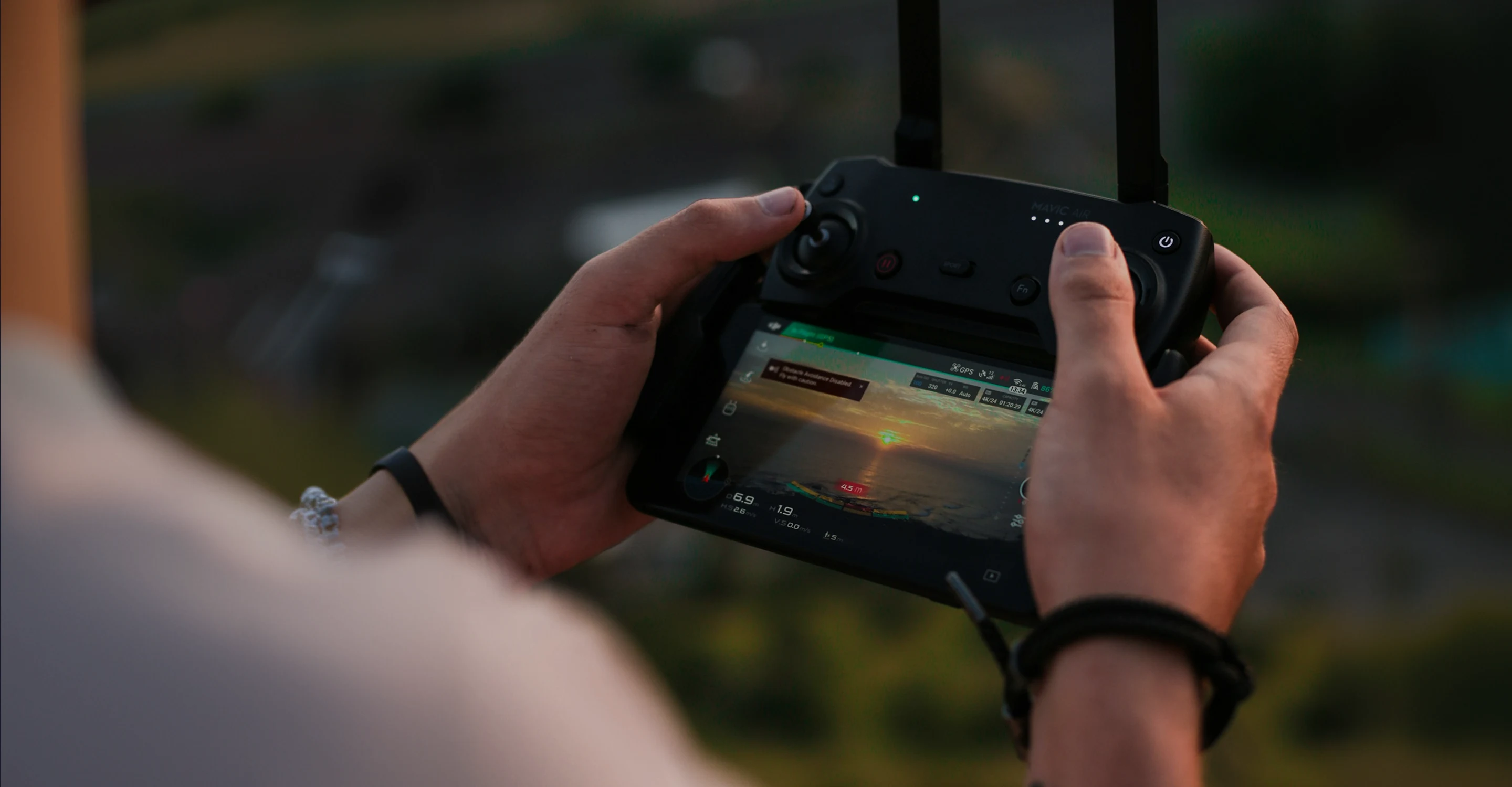 Person holding a drone remote controller with screen showing a sunset over water.