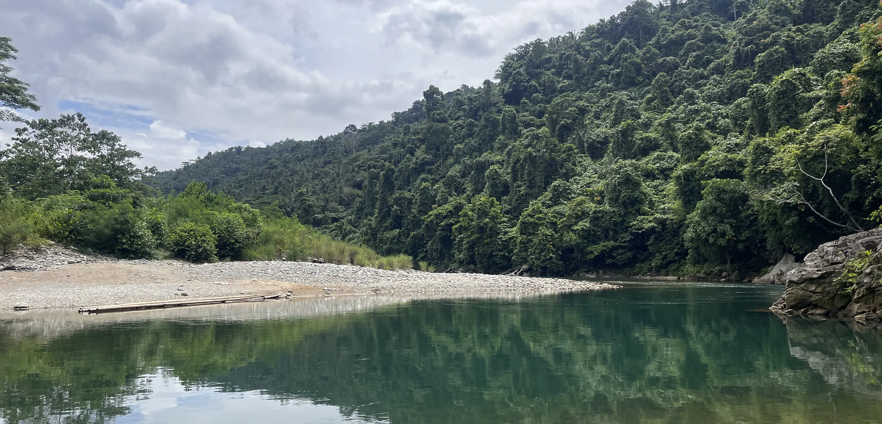 Calm river reflecting lush green forested hills under a partly cloudy sky.