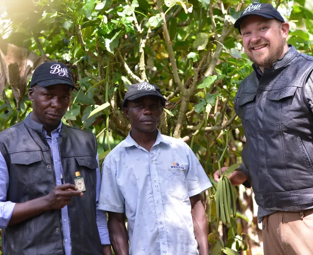 Three men in caps stand outdoors among green plants, one holding a small bottle, another holding green beans.