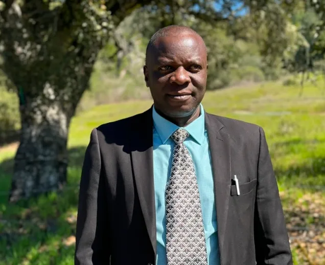 Man in a suit with a patterned tie standing outdoors with trees and grass in the background.