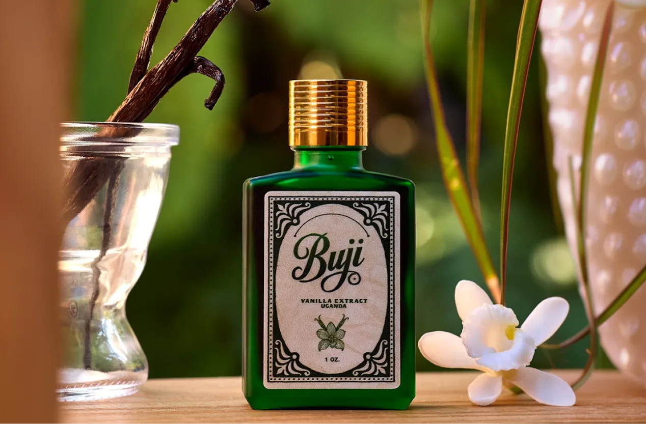 Green bottle of Buji Vanilla Extract Uganda with a vanilla flower and pods in glass on a wooden surface.