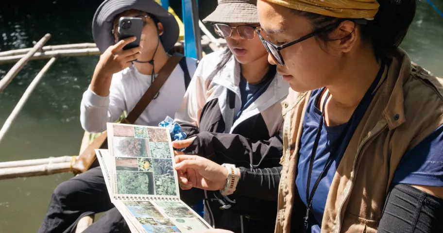 Three people on a boat examining a photo book with nature images under sunlight.